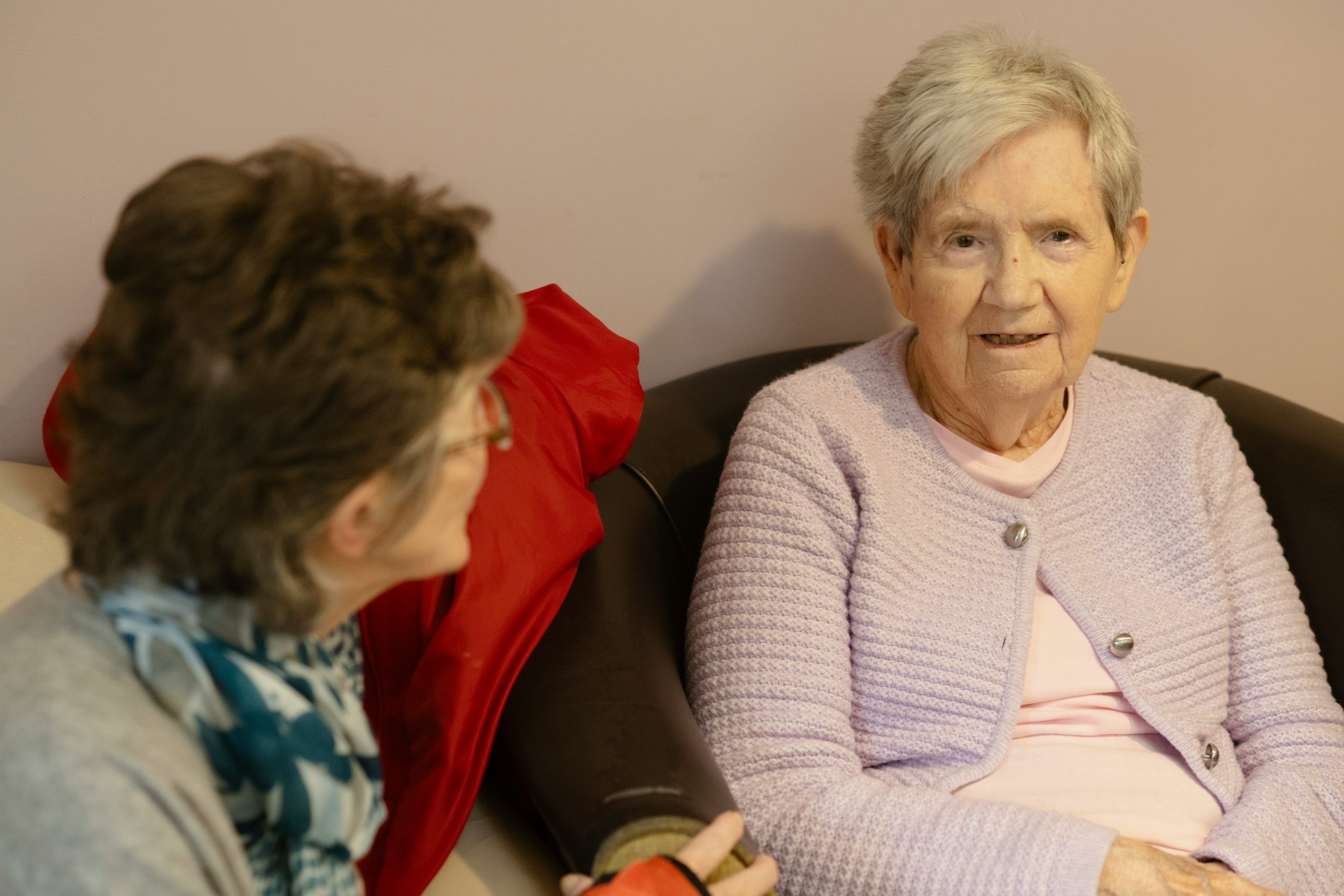 Seniorin sitzt im Gespräch mit einer Anghörigen auf dem Sofa und zeigt, wie persönliche Zuwendung die Grundpflege im Alltag ergänzt.