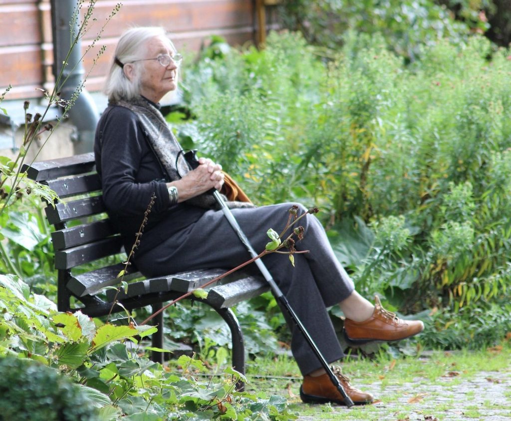 Ältere Frau sitzt mit Gehstock auf einer Bank im Grünen und genießt einen ruhigen Moment.