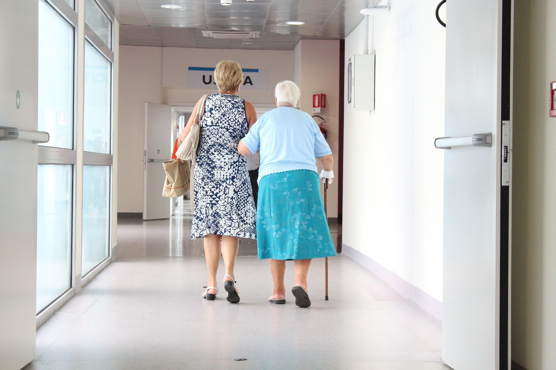 Begleitperson unterstützt eine ältere Frau mit Gehstock auf einem Krankenhausflur.